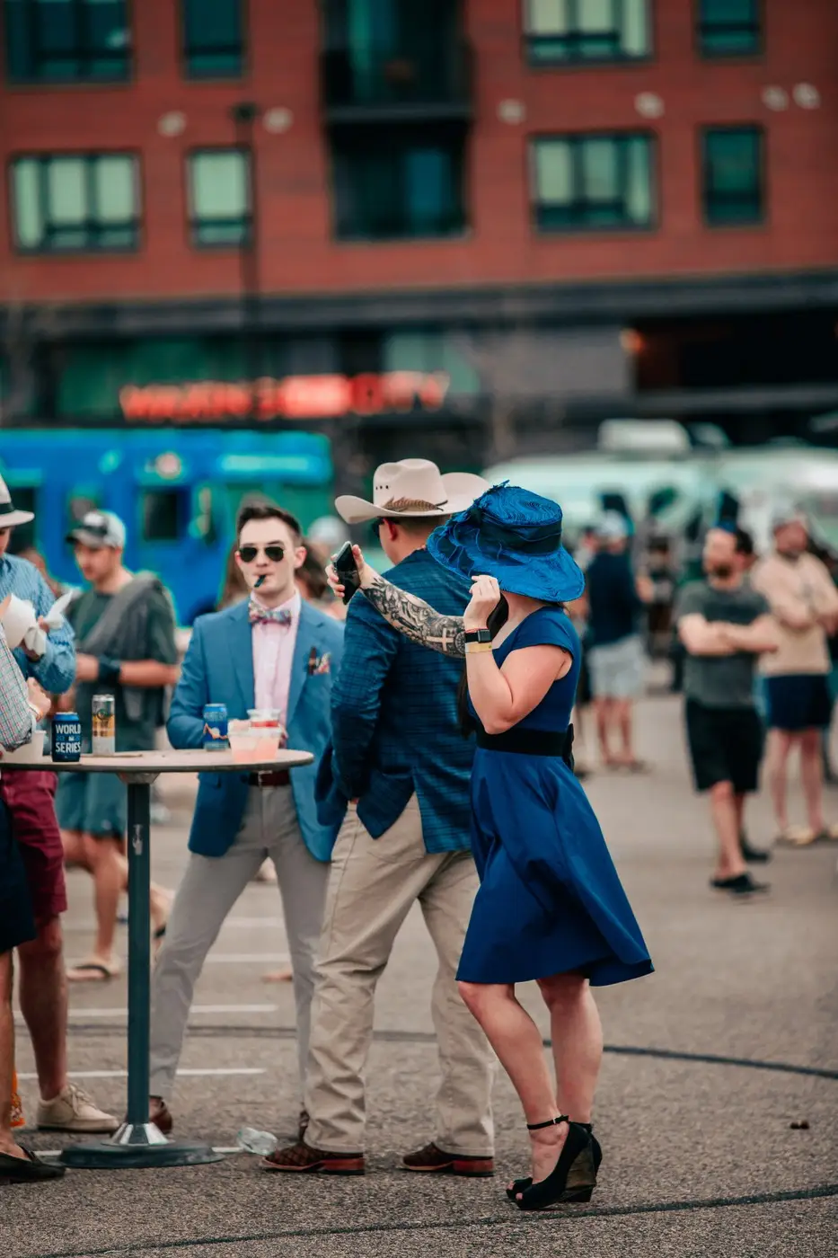 A couple in derby hats, him in a blue blazer, leaning in for a kiss.