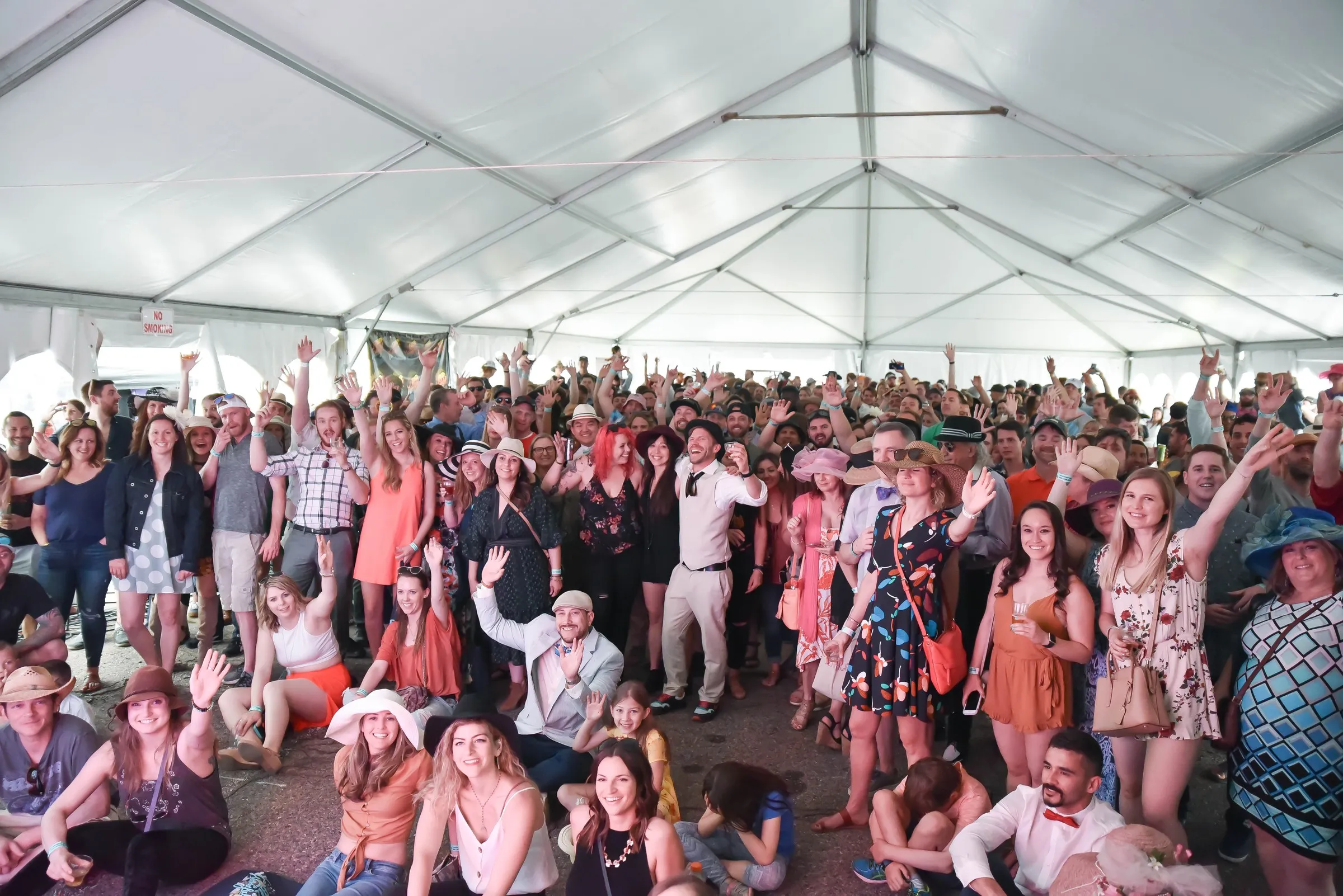 A packed tent of people in derby hats and spring dresses, all turned to the camera and cheering.