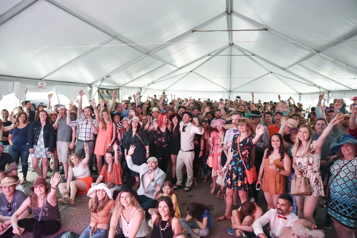 A packed tent of people in derby hats and spring dresses, all turned to the camera and cheering.
