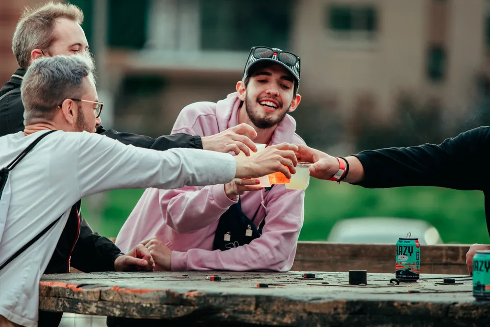 Two friends clinking cans across a wooden table at the RiNo festival