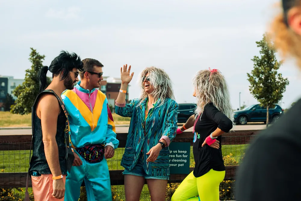 Four people in 80s wigs, a windbreaker tracksuit and neon leggings laughing together on the festival grounds