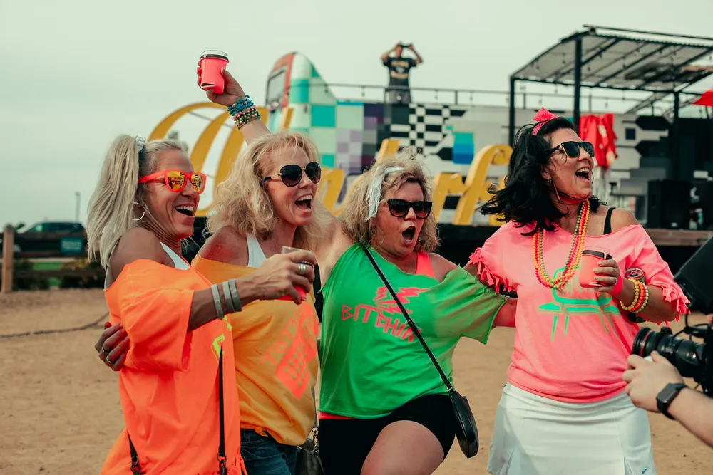 Four friends in neon 80s shirts, beads and sunglasses raising their cups in front of the big yellow Wonderful sign