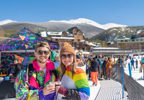 Two friends in rainbow ski suits with beers, mountain behind.